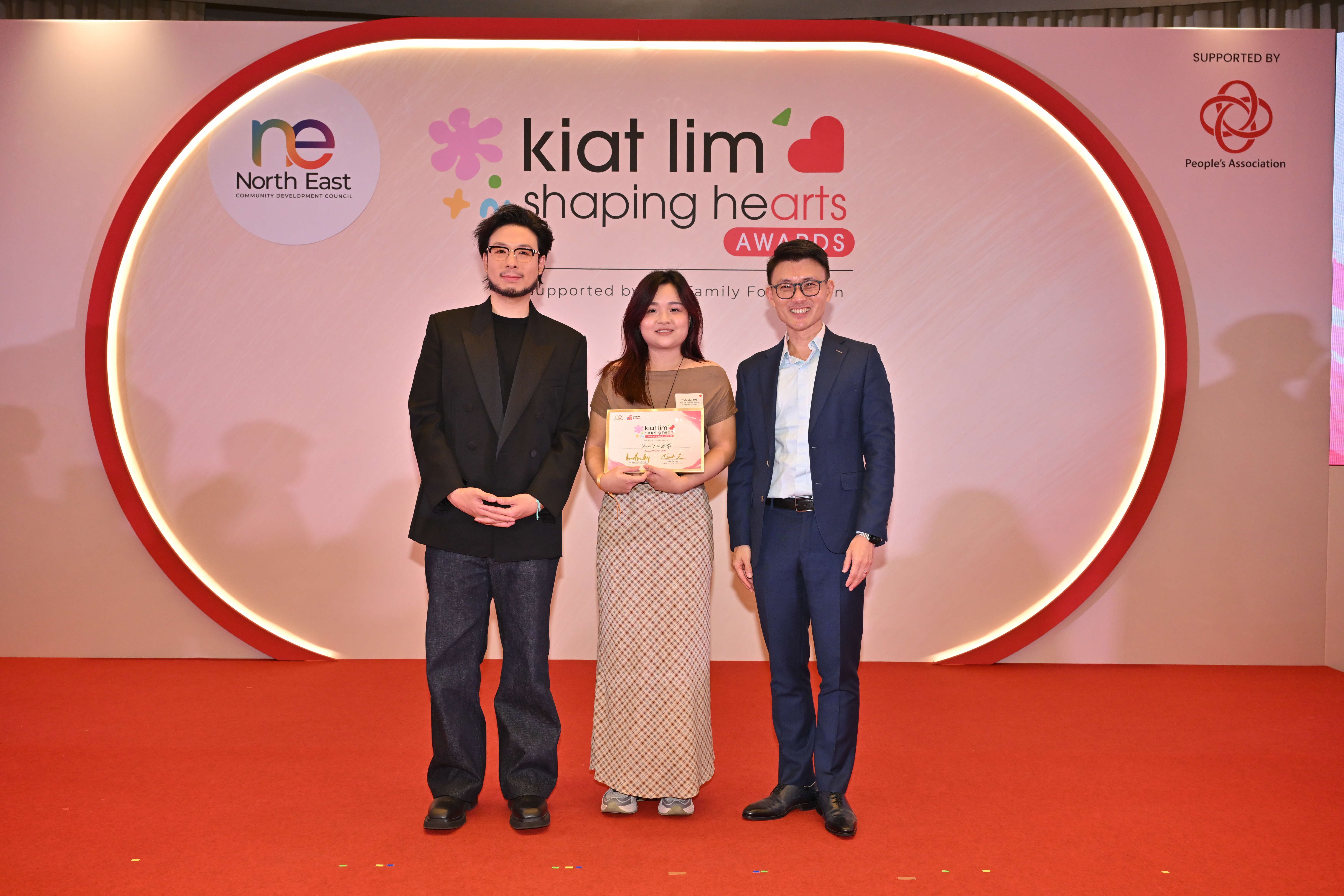 Award recipient standing on stage with the presenter, holding their certificate and posing for a group photo during the award ceremony, with a backdrop reading ‘Kiat Lim-Shaping Hearts Awards'.